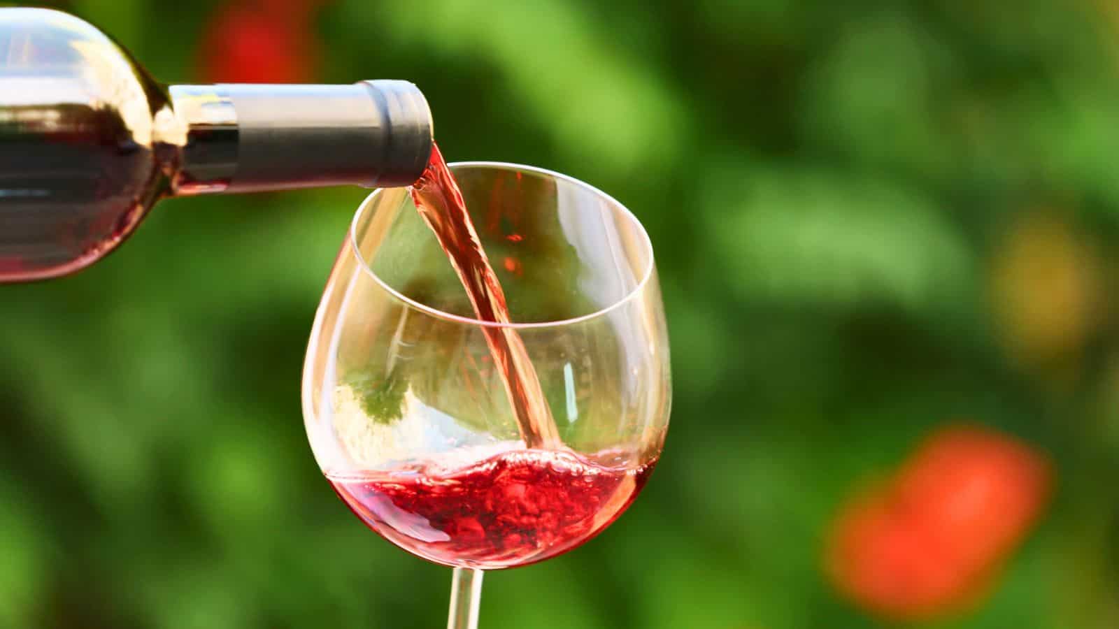 Red wine is being poured from a bottle into a wine glass against a blurred green and red background.