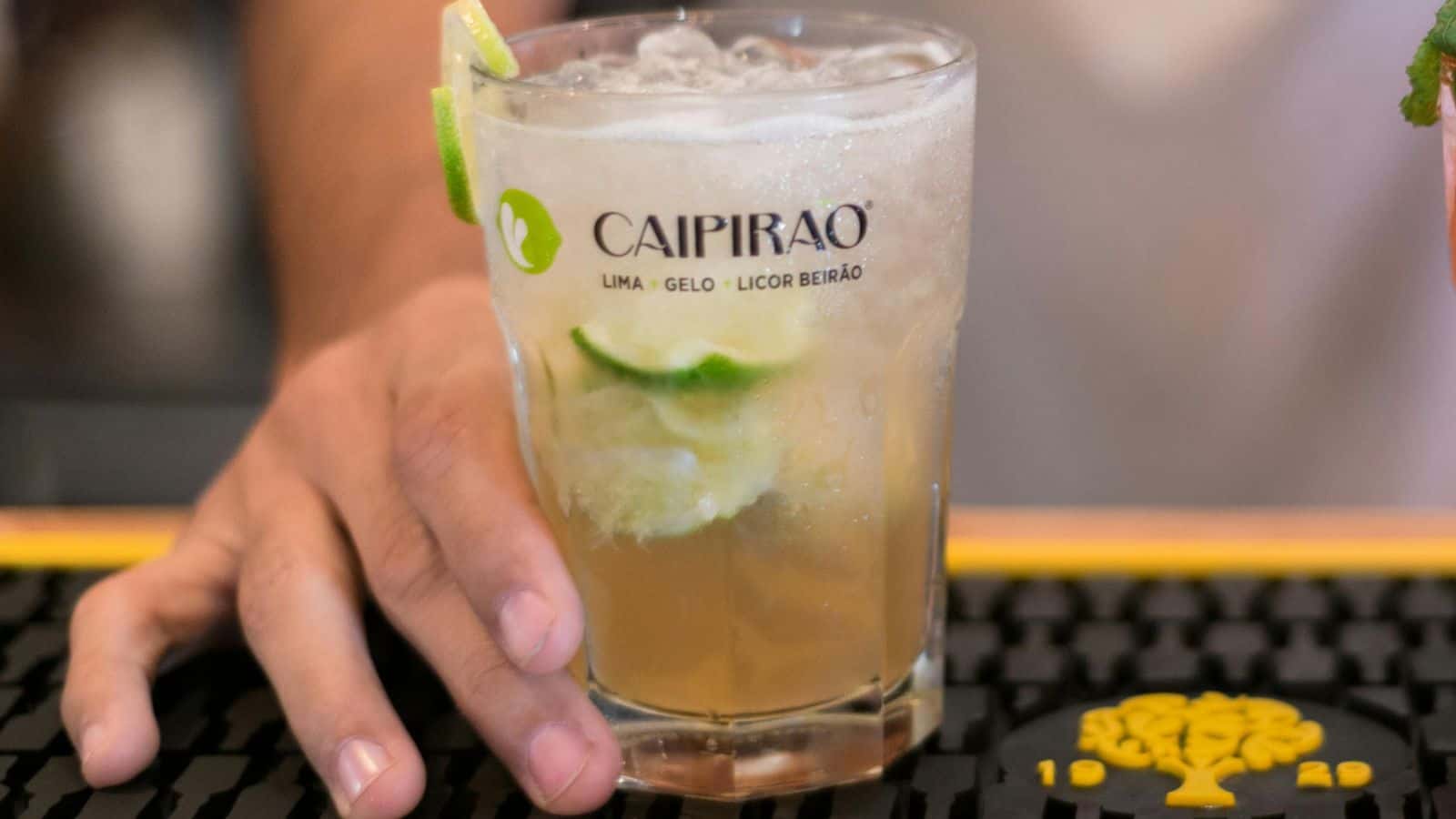 Close-up of a hand holding a glass of Caipirao, a cocktail containing lime, ice, and Licor Beir&atilde;o. The drink is garnished with lime, and a bar counter can be seen in the background.