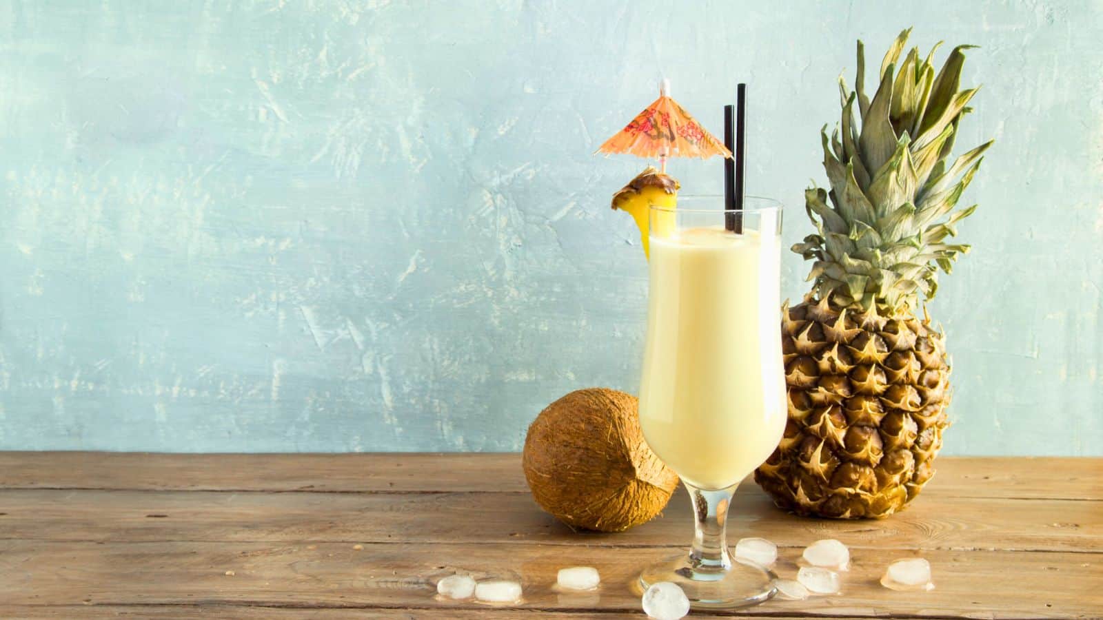 A glass of creamy pina colada with a slice of pineapple and a small umbrella sits on a wooden table. Beside it, there is a whole pineapple, a coconut, and scattered ice cubes against a light blue background.