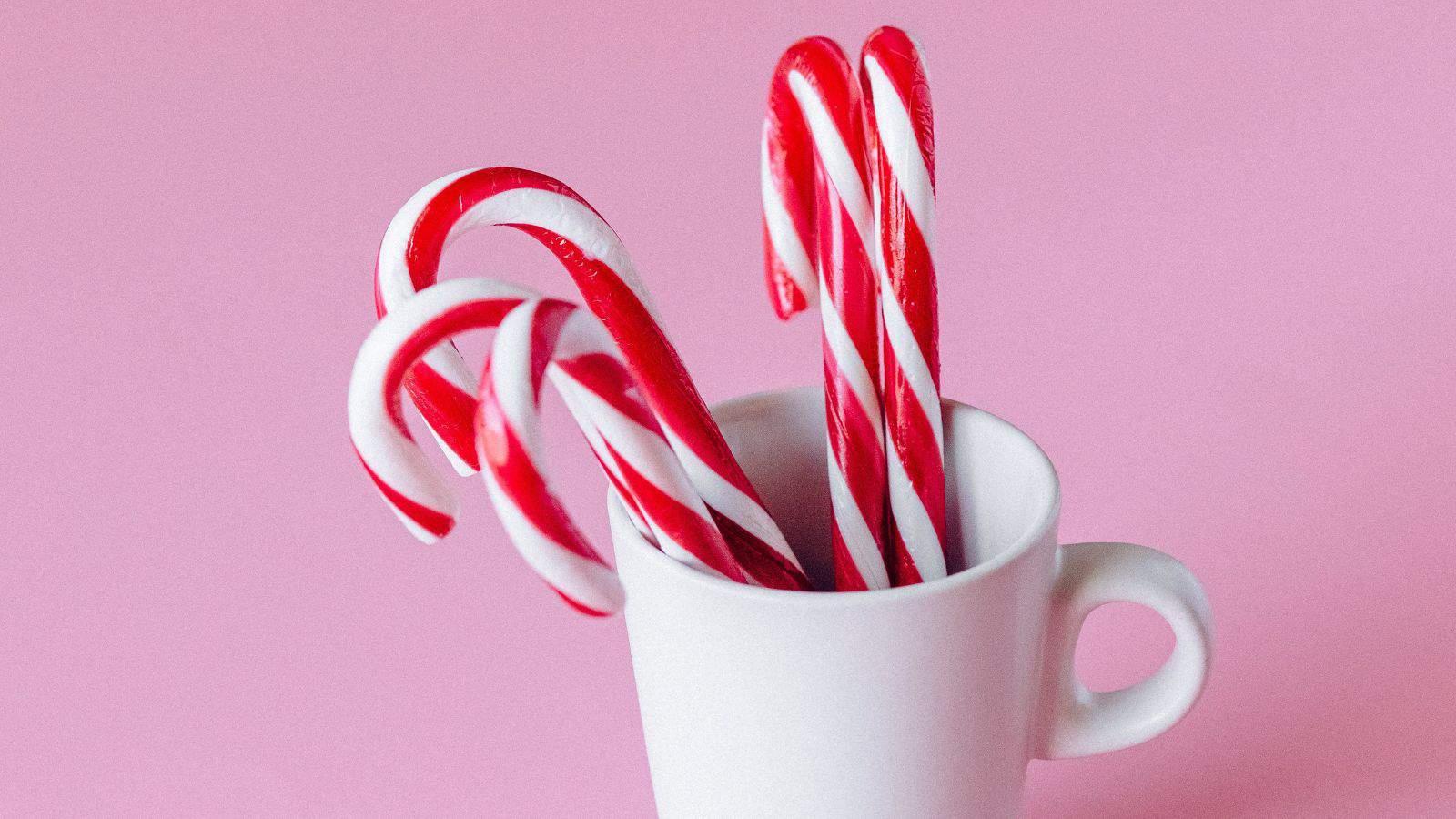 Three red and white striped candy canes are placed inside a white mug. The background is a solid pink color.