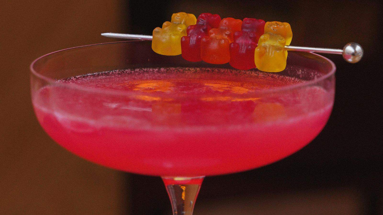 A vibrant pink cocktail in a glass garnished with a skewer of colorful gummy bears, adding a playful touch to the drink presentation.