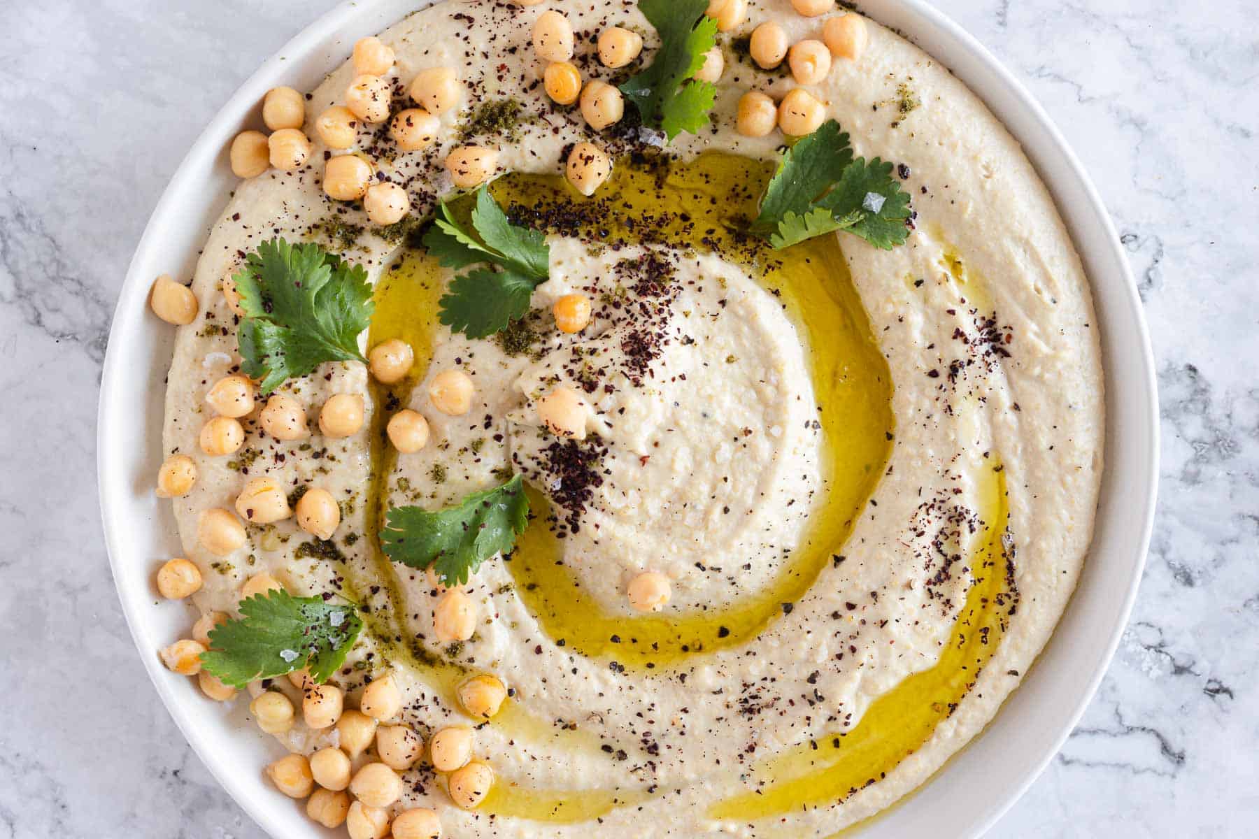 A bowl of hummus topped with chickpeas, olive oil, fresh cilantro, and sprinkled with spices. The dish is served on a marble surface.