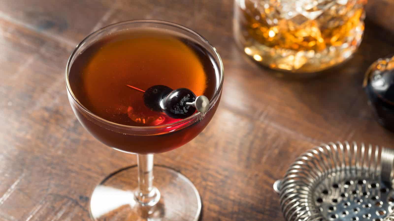 A close-up of a cocktail in a glass, garnished with two dark cherries on a cocktail pick. The drink is amber in color, set on a wooden surface. In the background are a cocktail shaker and a glass container with ice.