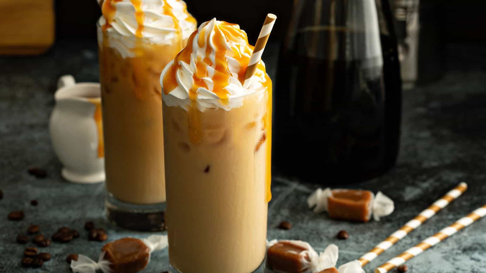 Two tall glasses of iced coffee topped with whipped cream and caramel drizzle, accompanied by striped straws. Caramel candies and coffee beans are scattered around the dark surface. A small jug and a dark bottle are in the background.
