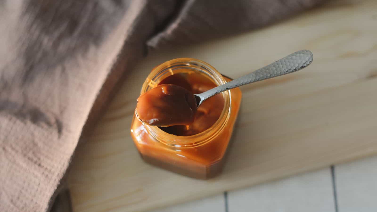 An open jar of caramel spread is placed on a wooden surface. A spoon rests across the top of the jar with a dollop of caramel on it. A textured brown cloth is partially visible in the background.