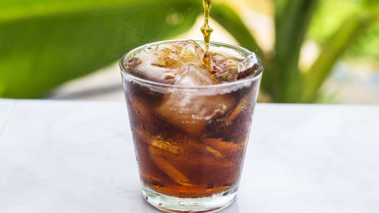 A glass filled with ice is placed on a table, with a dark liquid being poured into it. The background features a large green leaf out of focus.