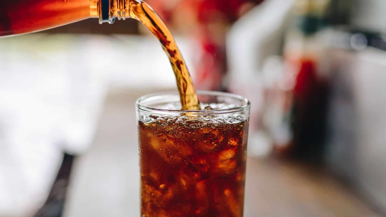 A glass is being filled with a fizzy brown liquid from a bottle. The glass contains ice cubes, and the liquid is pouring in, causing bubbles to form. The background is blurred, indicating an indoor setting.