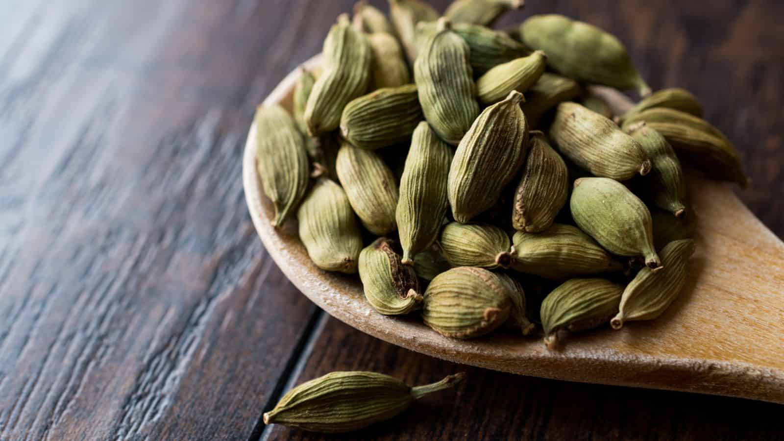 A wooden spoon holds a pile of green cardamom pods, resting on a dark wooden surface. One pod has fallen beside the spoon. The textured pods create a contrast with the smooth, rich background.