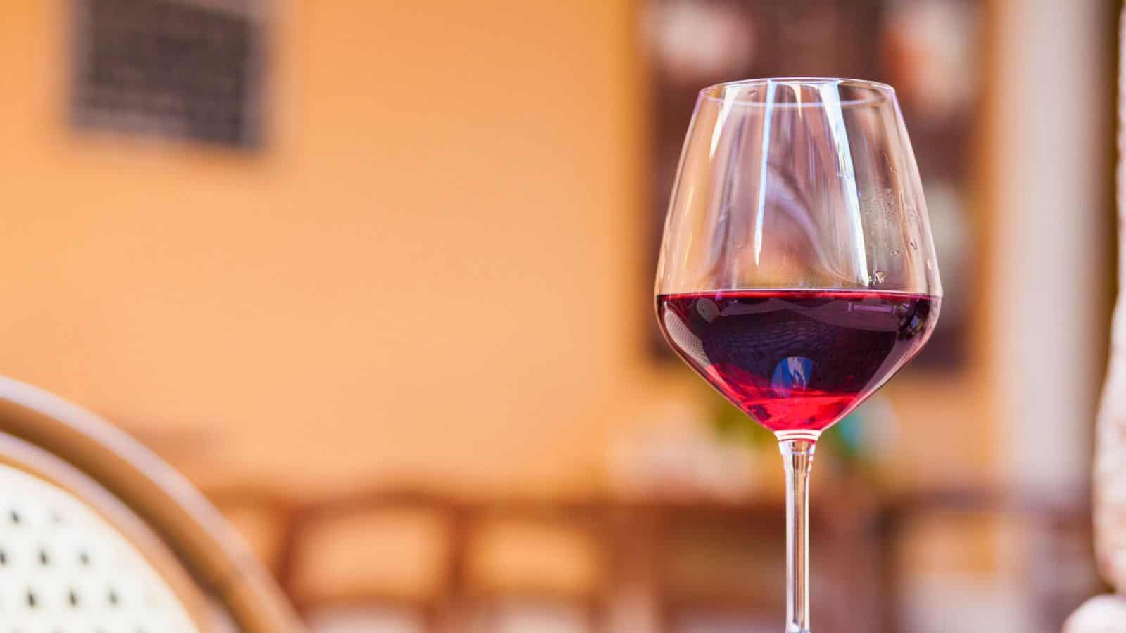 A glass of red wine is placed on an outdoor table with a blurred background of chairs and an orange wall, suggesting an outdoor cafe or patio setting.