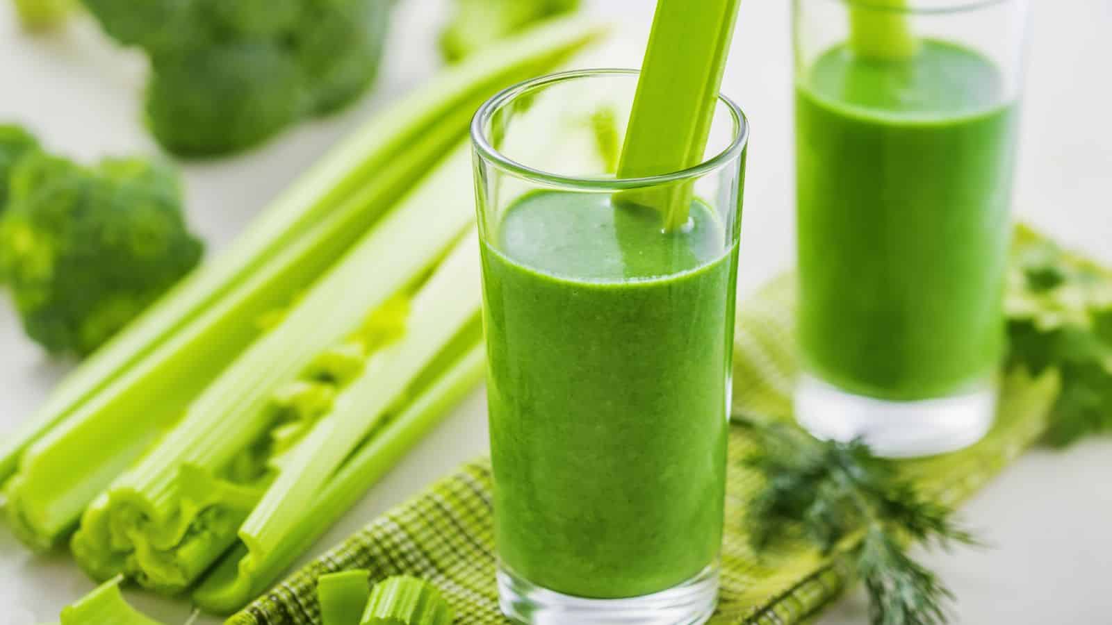 Two glasses filled with green smoothie are on a checkered cloth. Each glass has a celery stalk. Surrounding them are fresh celery stalks and broccoli florets. The focus is on the vibrant green color of the smoothies and vegetables.