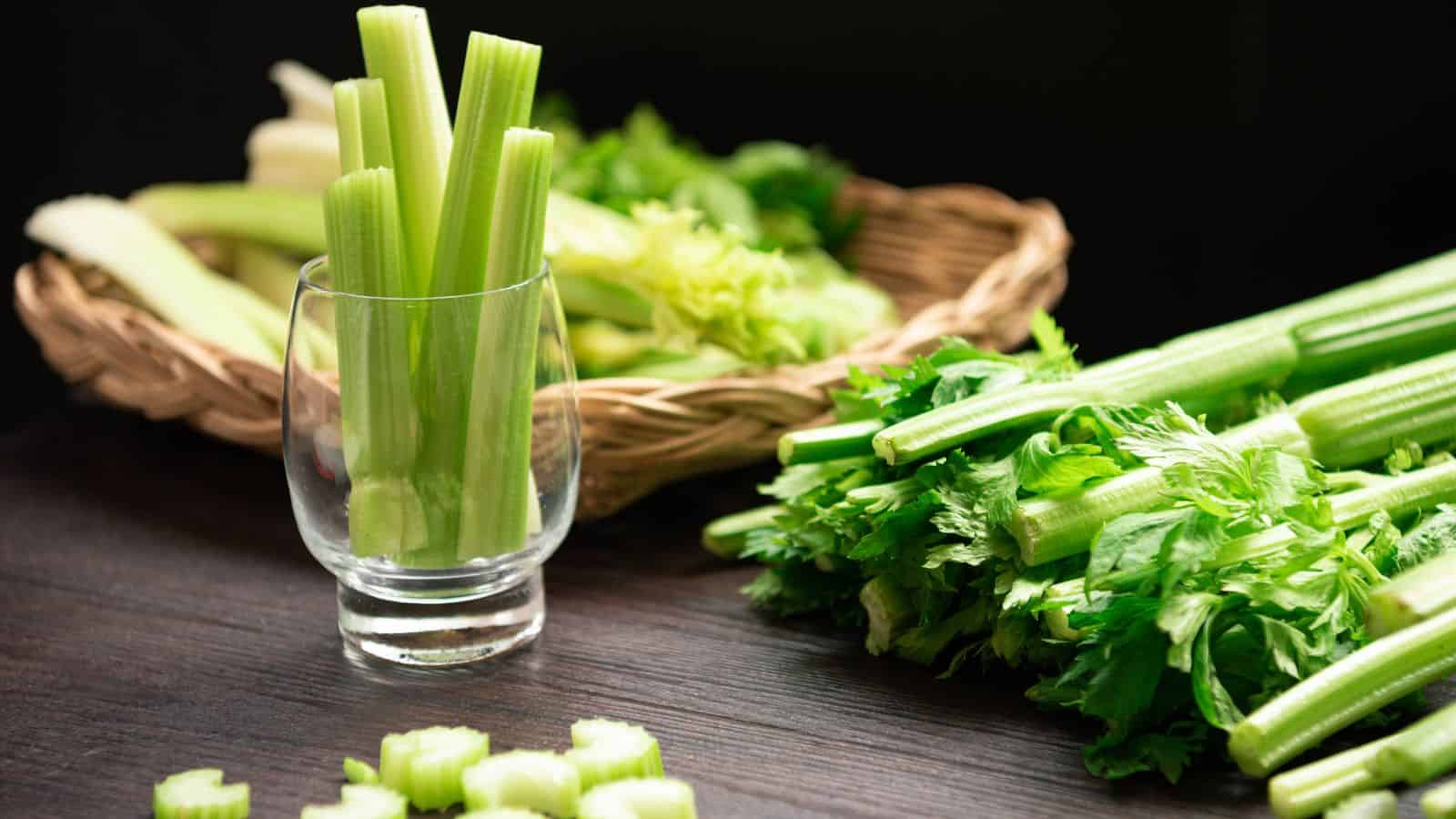 A glass filled with upright celery stalks is in the foreground on a dark wooden surface. Behind it, a basket holds more celery. Additional celery stalks and leaves are scattered to the side, with some cut pieces on the table.