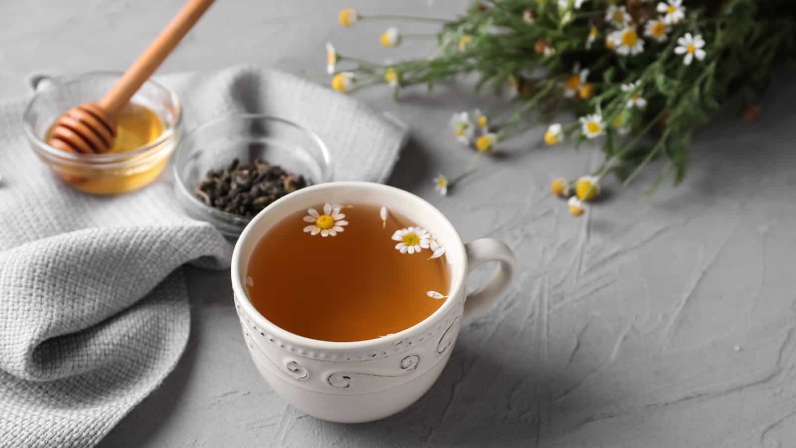 A cup of chamomile tea sits on a textured gray surface. Nearby are a small bowl of honey with a dipper and a bowl of loose tea leaves on a folded gray cloth. A bunch of chamomile flowers lies on the side.