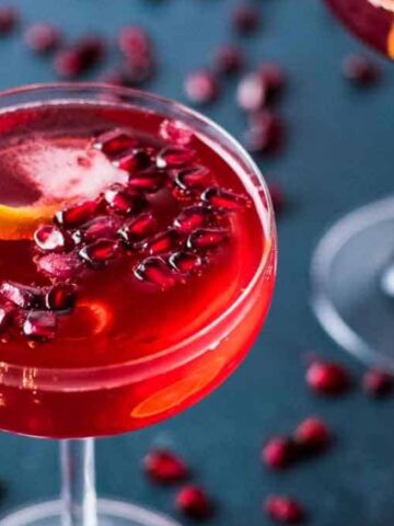 A close-up of a red cocktail in a glass, garnished with pomegranate seeds and an orange peel twist. Another similar glass is partially visible in the background on a dark surface. Pomegranate seeds are scattered around the glasses.