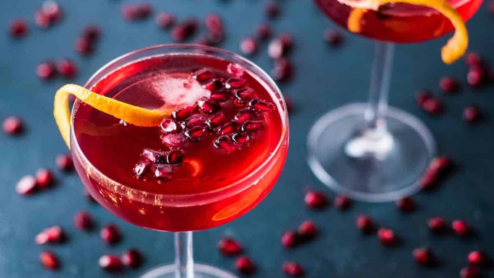 A close-up of a red cocktail in a glass, garnished with pomegranate seeds and an orange peel twist. Another similar glass is partially visible in the background on a dark surface. Pomegranate seeds are scattered around the glasses.