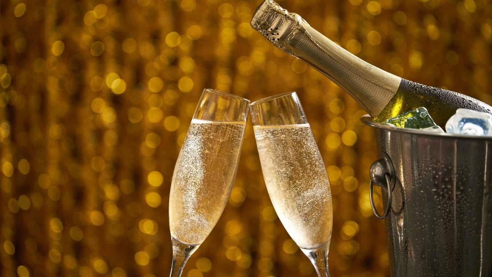 Two champagne glasses filled with bubbly liquid clink together in front of a golden, bokeh background. A champagne bottle rests in an ice bucket on the right, adding to the celebratory atmosphere.