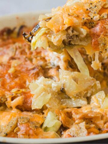A close-up of a cheesy casserole with golden-brown crust in a white baking dish. One portion is being lifted, showing melted cheese and crunchy toppings.
