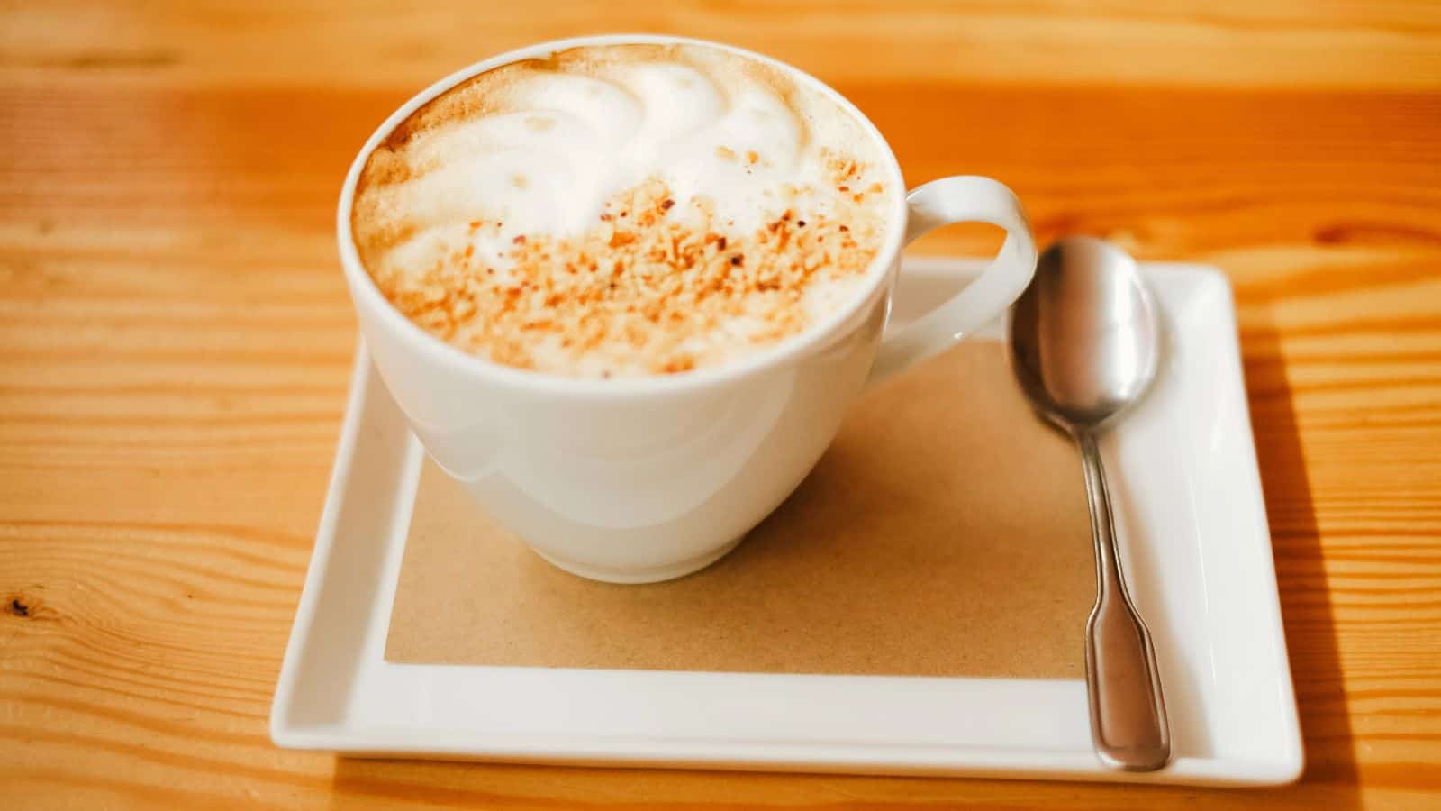 A cup of cappuccino with frothy milk and a sprinkle of cocoa sits on a white square saucer. A silver spoon rests beside it. The table underneath is made of light-colored wood.