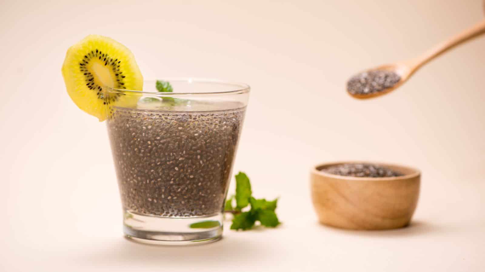 A glass of chia seed pudding garnished with a slice of kiwi and mint leaves is placed on a light surface. A wooden spoon with chia seeds is held above a small wooden bowl, partially visible in the background.