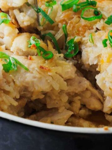 A casserole dish filled with a baked chicken and rice dish, topped with melted cheese and garnished with chopped green onions. A slice is missing from the dish, revealing the creamy texture inside.