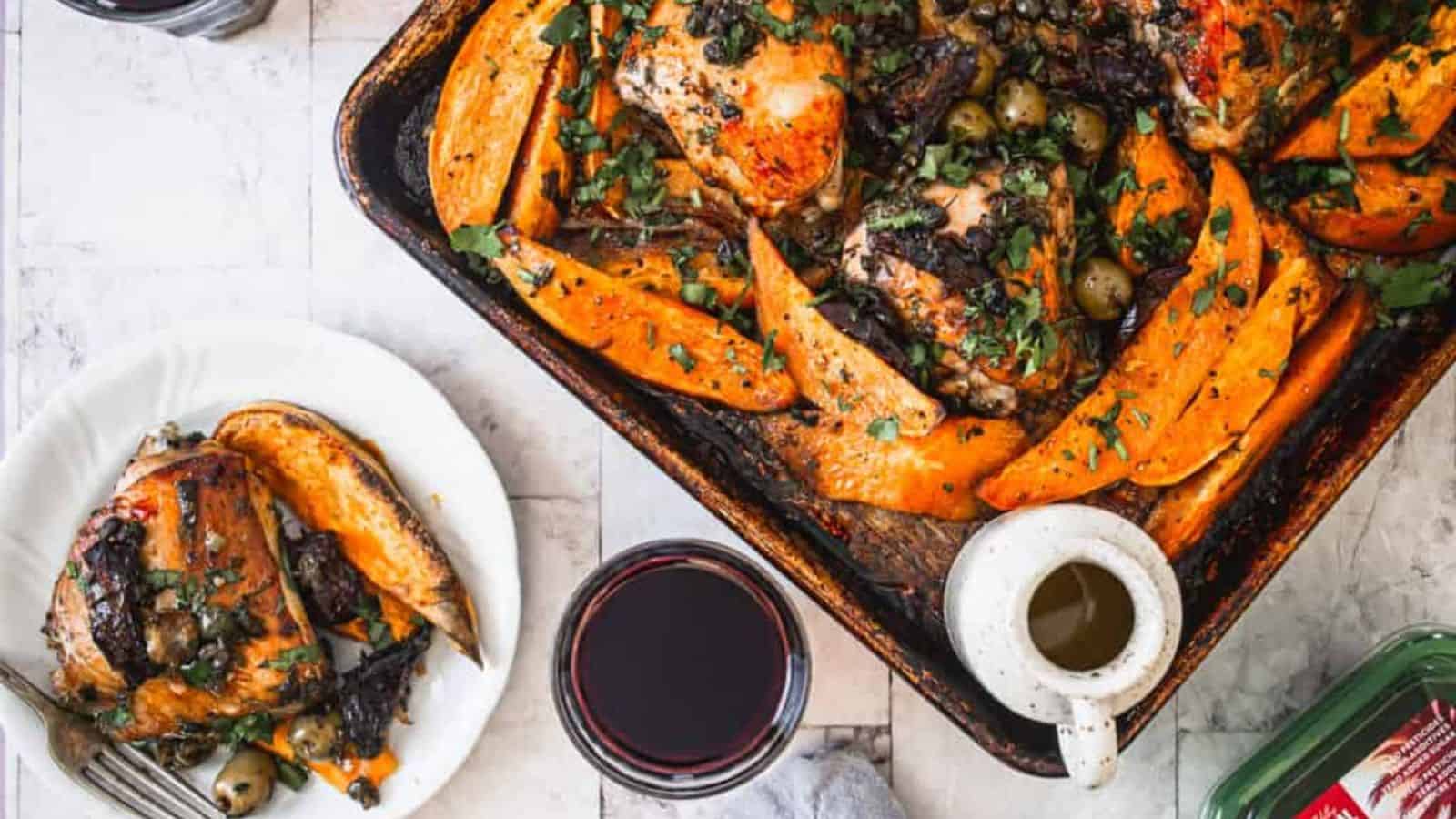 A roasted chicken and sweet potato dish is served in a baking tray, garnished with herbs and olives. A serving of the dish is placed on a plate next to a glass of red wine and a small jug of sauce on a light-colored surface.