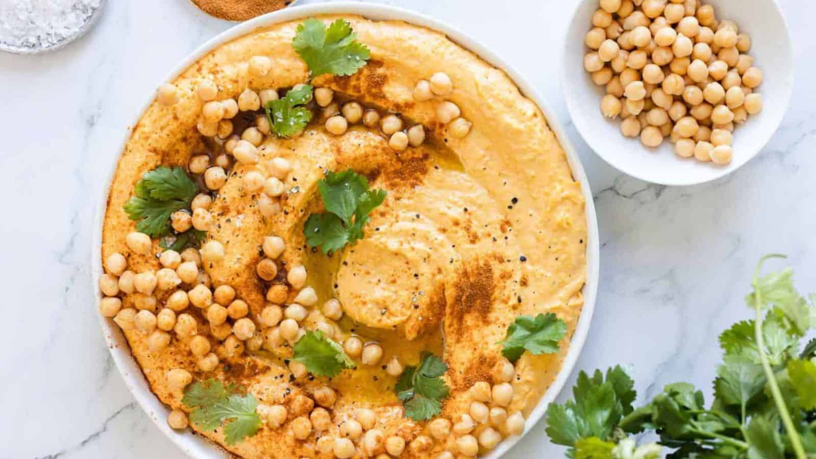 A bowl of hummus is topped with chickpeas, cilantro, and paprika. Nearby, a small dish contains extra chickpeas. A sprig of cilantro decorates the bottom corner of the image, which is set on a white marble surface.