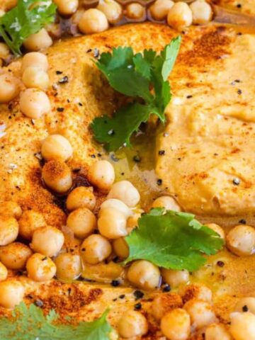 A close-up of a bowl of hummus topped with chickpeas, olive oil, paprika, black pepper, and cilantro leaves. The hummus has a creamy texture and is visually garnished with spices and herbs.