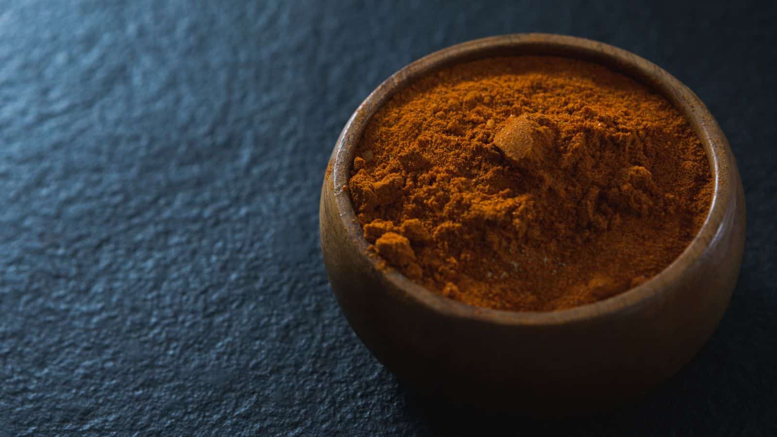 A wooden bowl filled with orange-brown powder, possibly a type of spice, sits on a dark textured surface. The lighting highlights the texture of the powder.