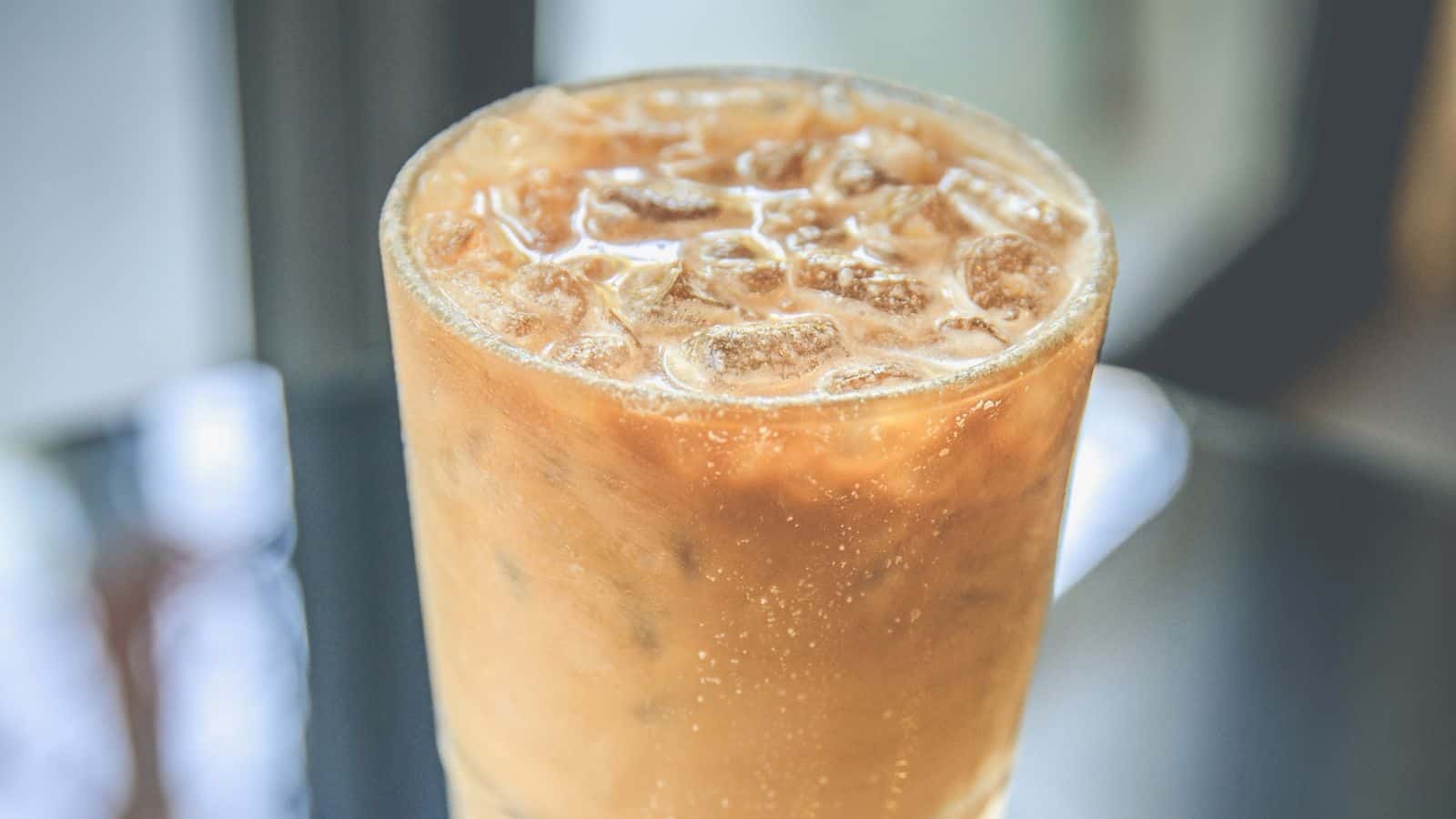 A tall glass filled with iced coffee, showing a light brown color, sits on a reflective surface. The coffee is topped with ice cubes. The background is softly blurred, suggesting an indoor setting with natural light.