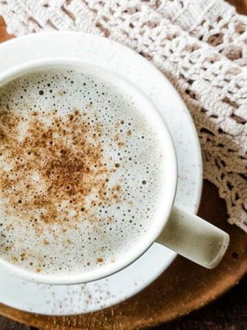 A white cup filled with a frothy beverage, dusted with cinnamon, sits on a white saucer. It is placed on a wooden tray next to a few star anise and a crocheted cloth. The background features a rustic wooden surface.