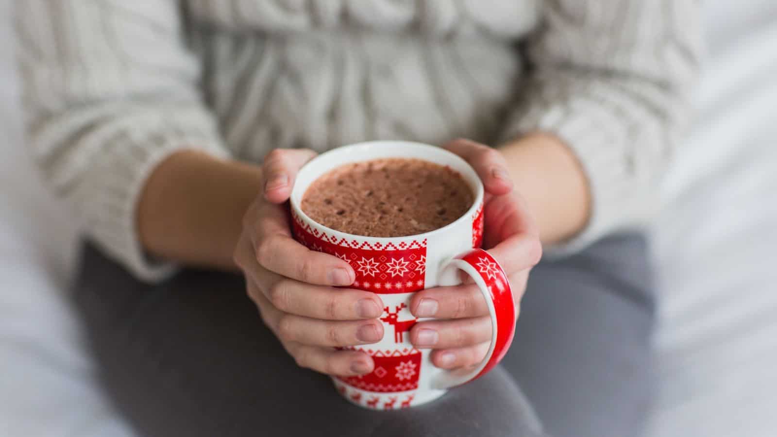 A person holding a red and white patterned mug filled with hot chocolate. The person is wearing a knitted sweater and sitting cross-legged on a soft surface. The mug features snowflakes and reindeer designs.