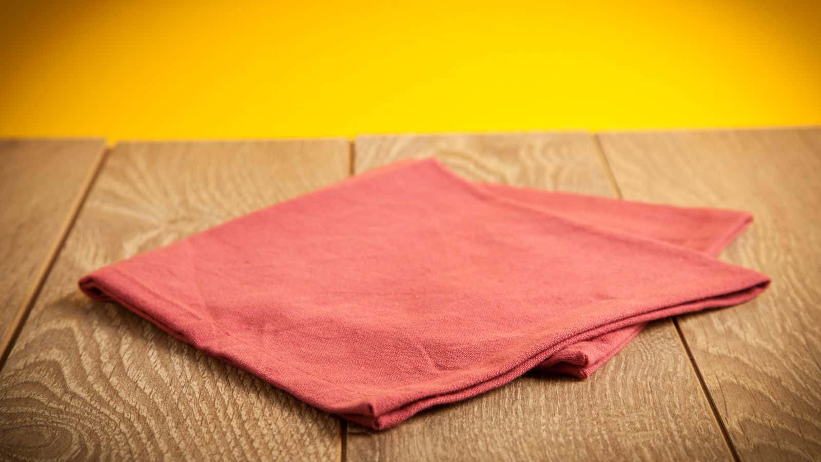 A pink cloth is neatly folded on a wooden surface with a bright yellow background.