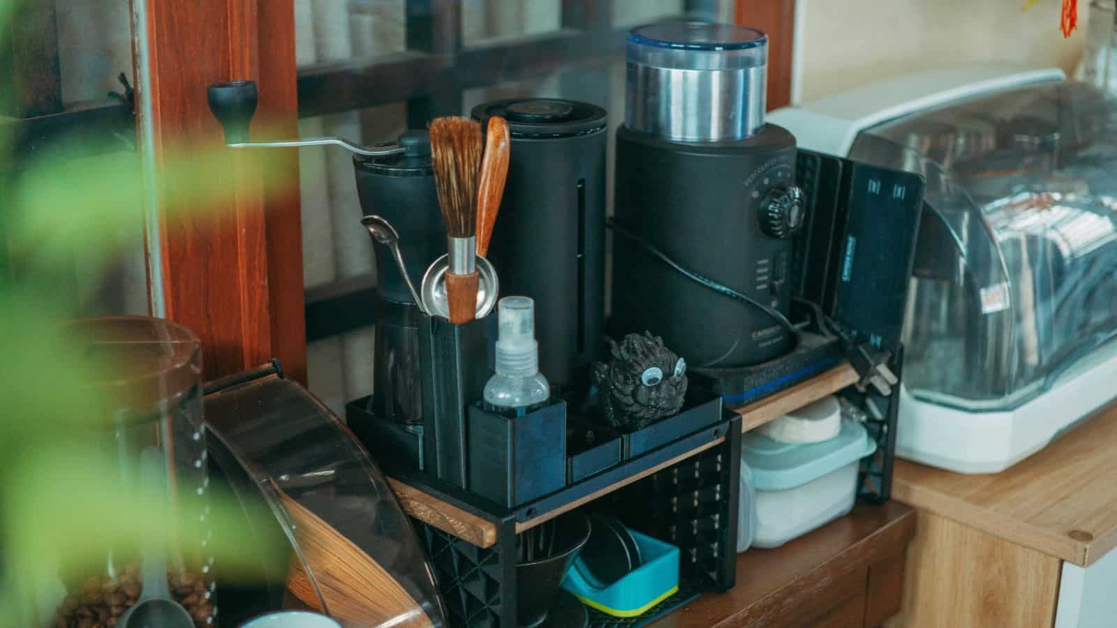 A kitchen countertop with various appliances, including a coffee grinder, a coffee maker, a breadbox, and a small spray bottle. There is a plant partially visible in the foreground.