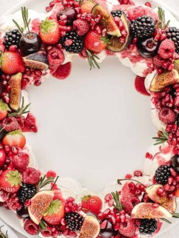 A festive wreath-shaped dessert on a white plate, topped with various fruits including strawberries, cherries, blackberries, raspberries, figs, and pomegranate seeds, garnished with sprigs of rosemary.