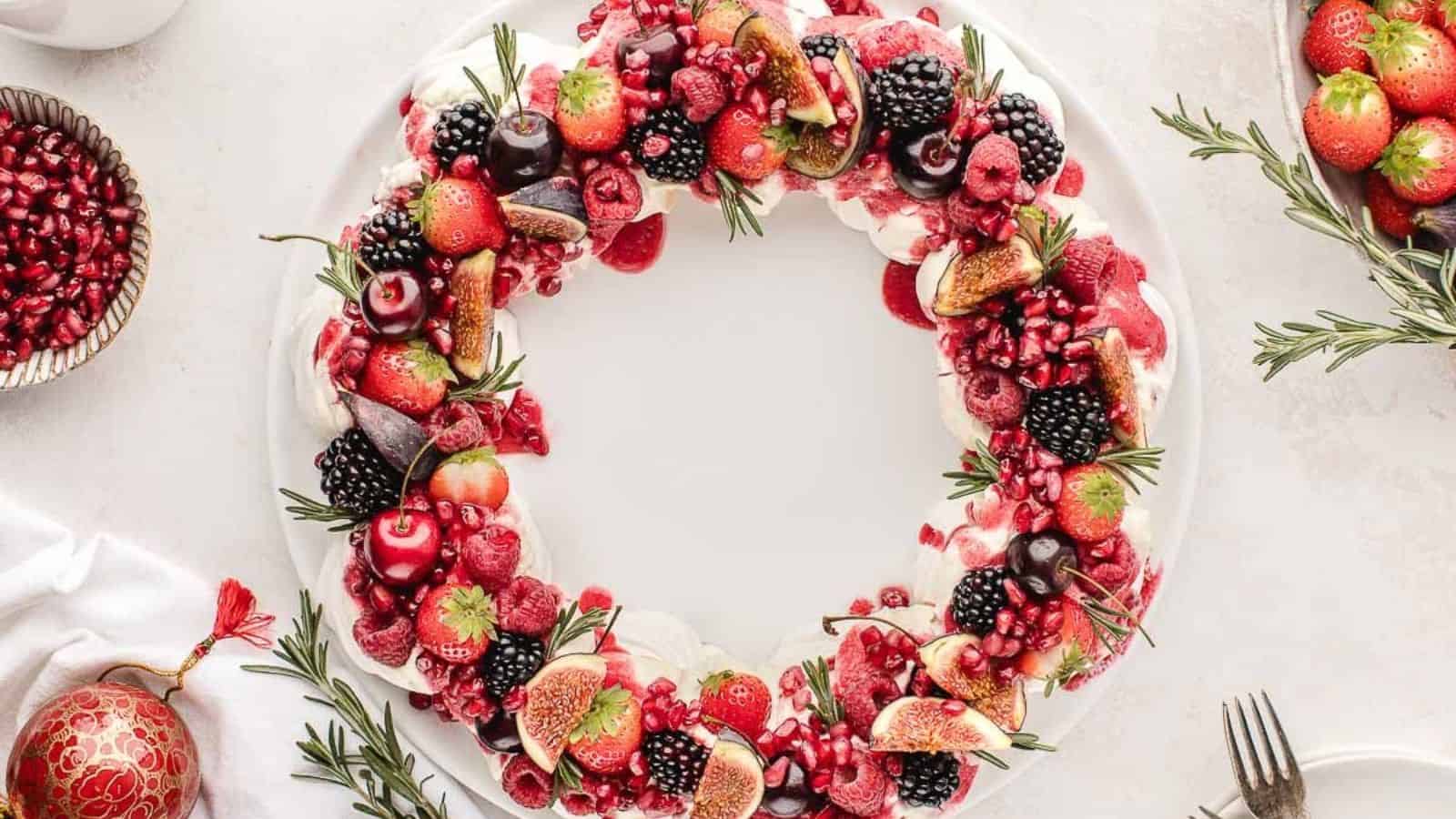 A festive wreath-shaped dessert on a white plate, topped with various fruits including strawberries, cherries, blackberries, raspberries, figs, and pomegranate seeds, garnished with sprigs of rosemary.