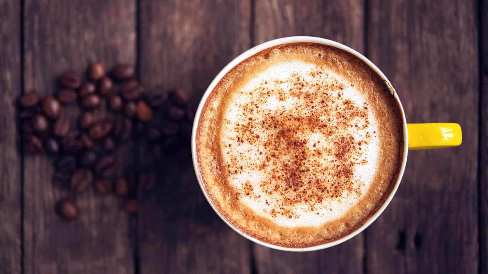A yellow cup filled with a frothy cappuccino topped with a sprinkle of cocoa or cinnamon sits on a wooden surface. Coffee beans are scattered to the left of the cup.