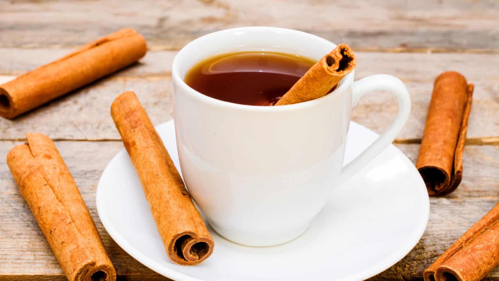 A white cup filled with tea sits on a saucer on a wooden surface. A cinnamon stick is placed in the cup, and several cinnamon sticks are scattered around the saucer.