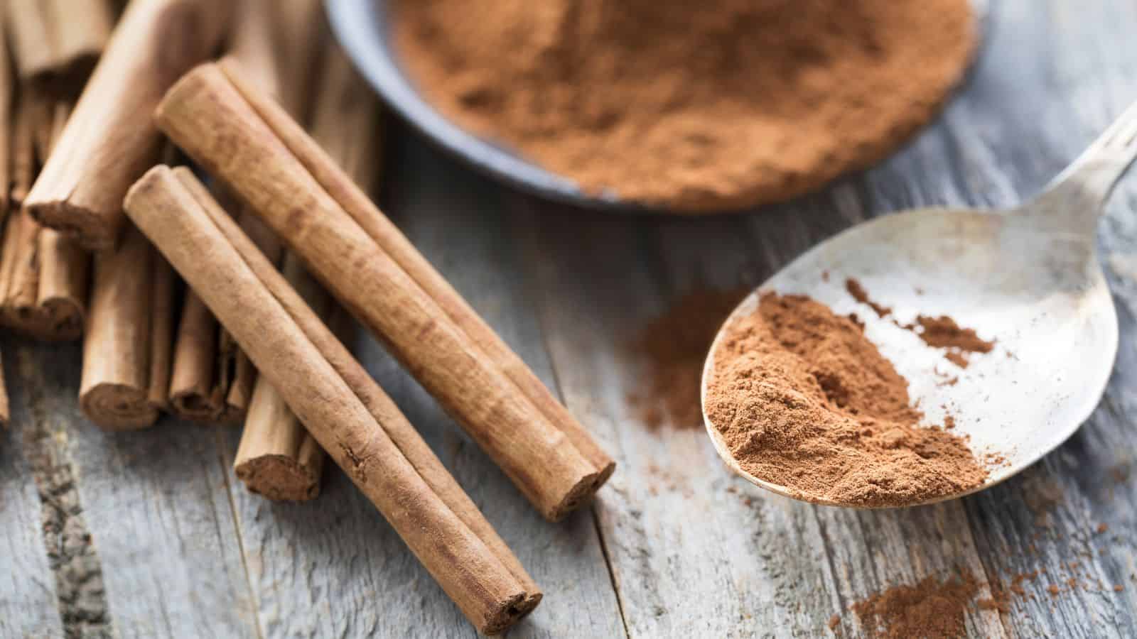 A close-up image of three cinnamon sticks on a wooden surface, next to a spoon filled with ground cinnamon. In the background, a plate with more ground cinnamon is partially visible. The scene captures a warm, rustic atmosphere.
