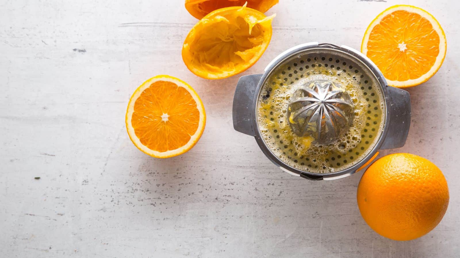 A metal citrus juicer with orange pulp sits surrounded by two squeezed orange halves and one whole orange on a white textured surface.