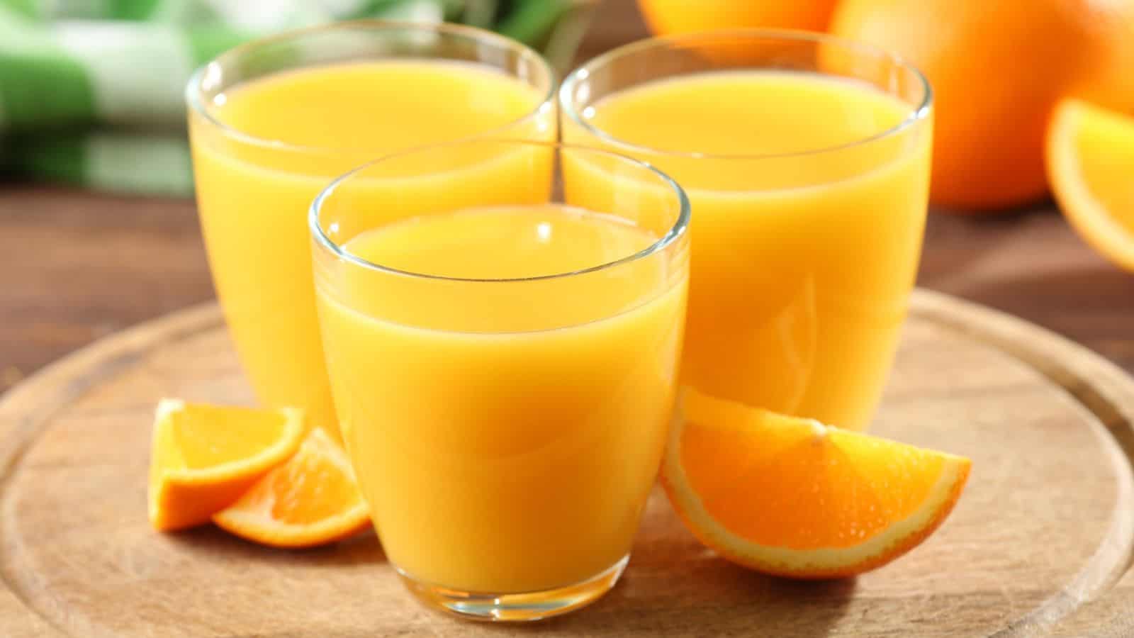 Three glasses of orange juice are on a wooden tray with orange slices beside them. Whole oranges are visible in the background.