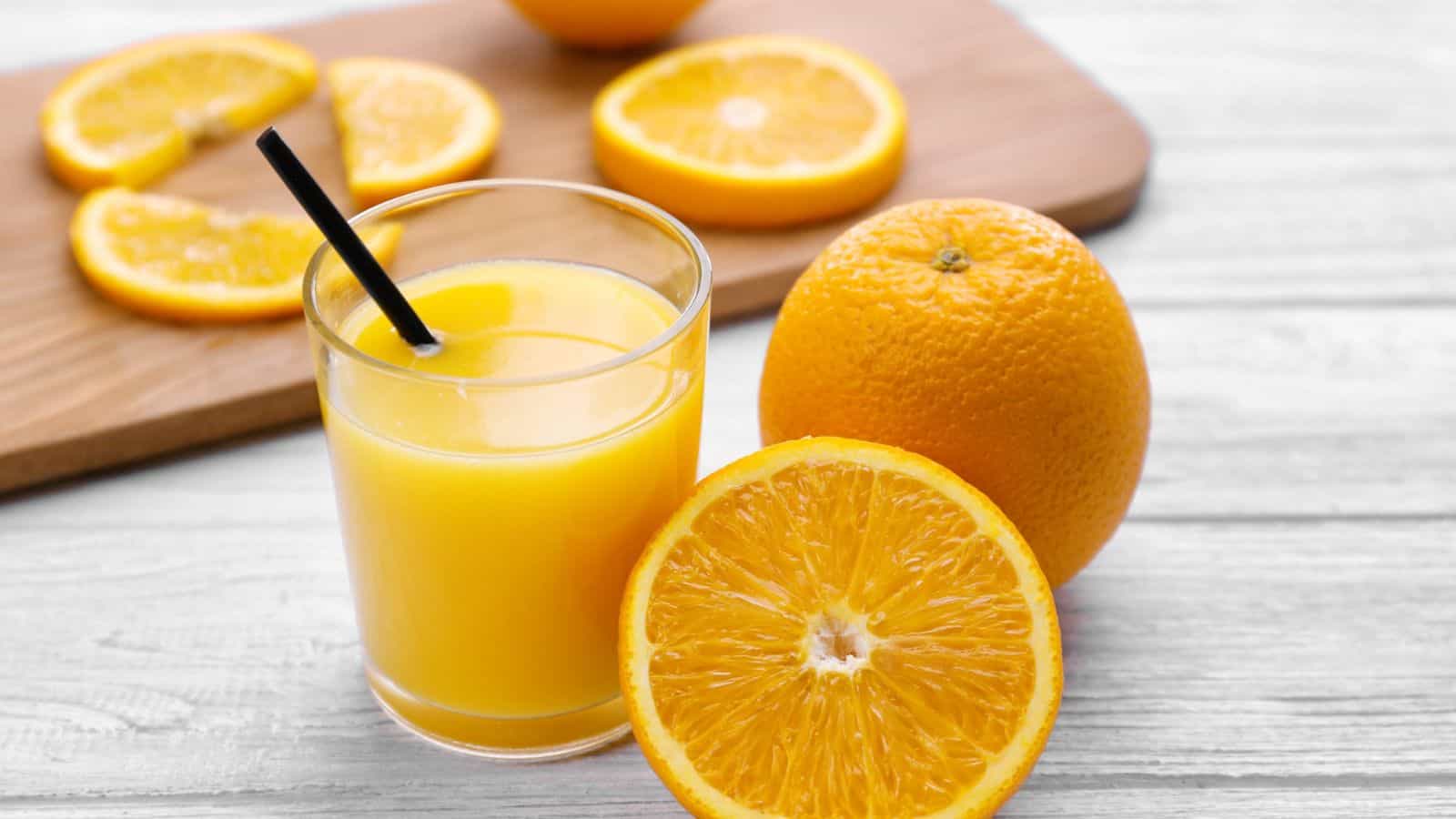 A glass of orange juice with a black straw sits on a white wooden surface. Beside it is a whole orange and a halved orange. In the background, orange slices are displayed on a wooden cutting board.