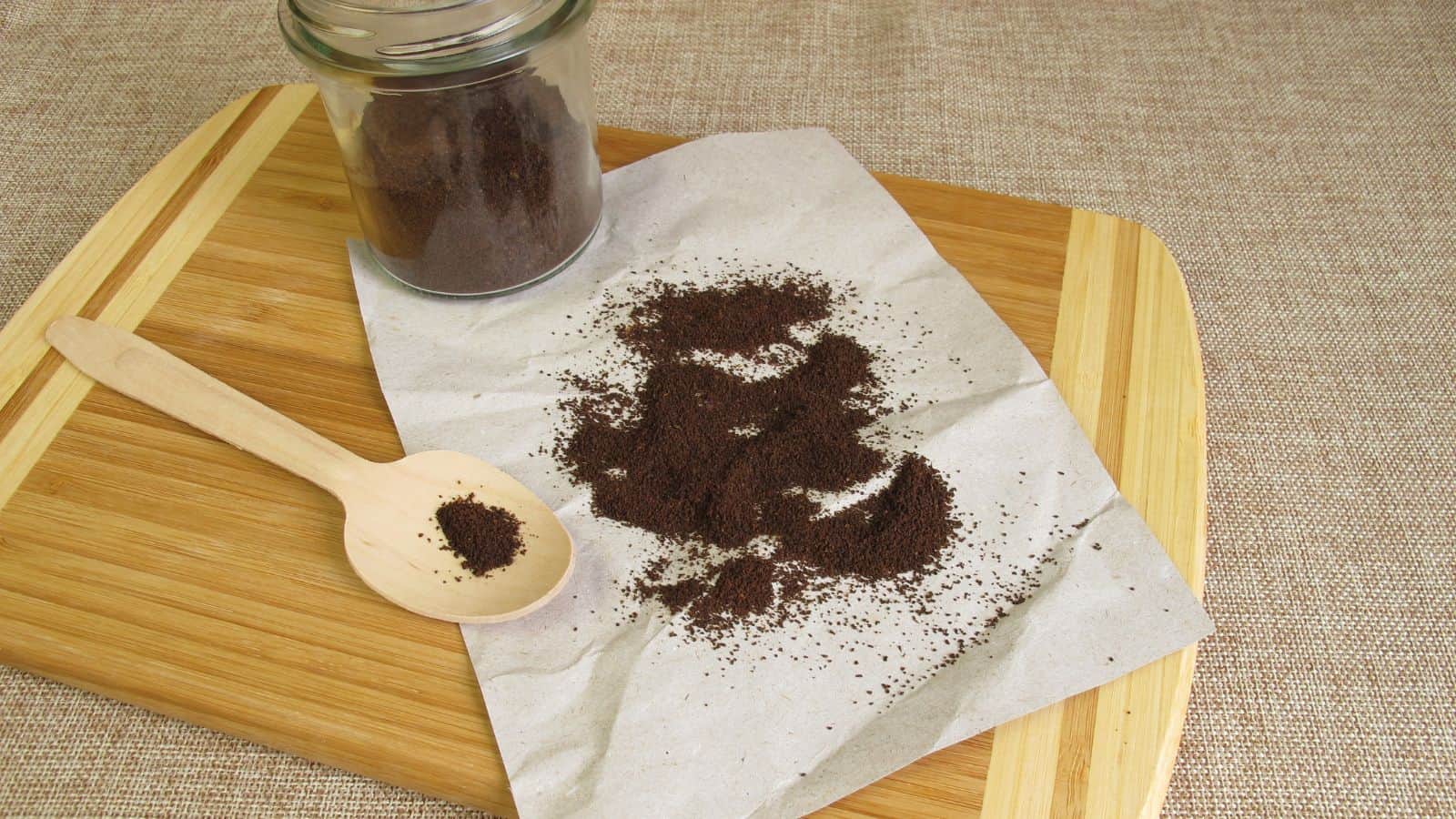 A glass jar tipped over, spilling ground coffee onto a sheet of white paper on a wooden cutting board. A wooden spoon with ground coffee on it lies nearby. The background surface is a textured beige fabric.
