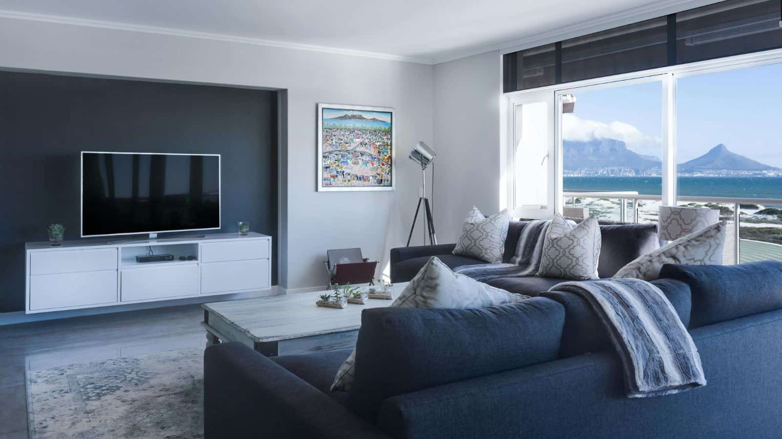 Modern living room with a large navy sofa, a light-colored coffee table, and a wall-mounted flat-screen TV. There is a painting on the wall and a tripod lamp. Large windows offer views of a mountainous landscape and the sea.