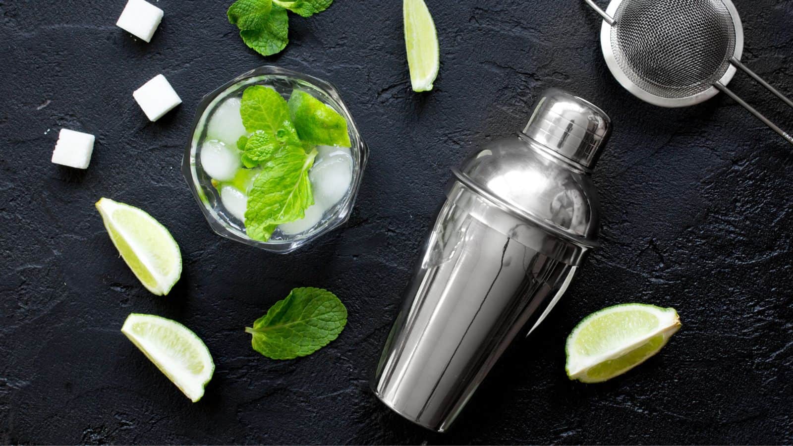 A cocktail shaker, cocktail strainer, and a glass filled with a green drink garnished with mint leaves are on a black surface. Ice cubes and lime wedges are scattered around. Sugar cubes are positioned near the glass.
