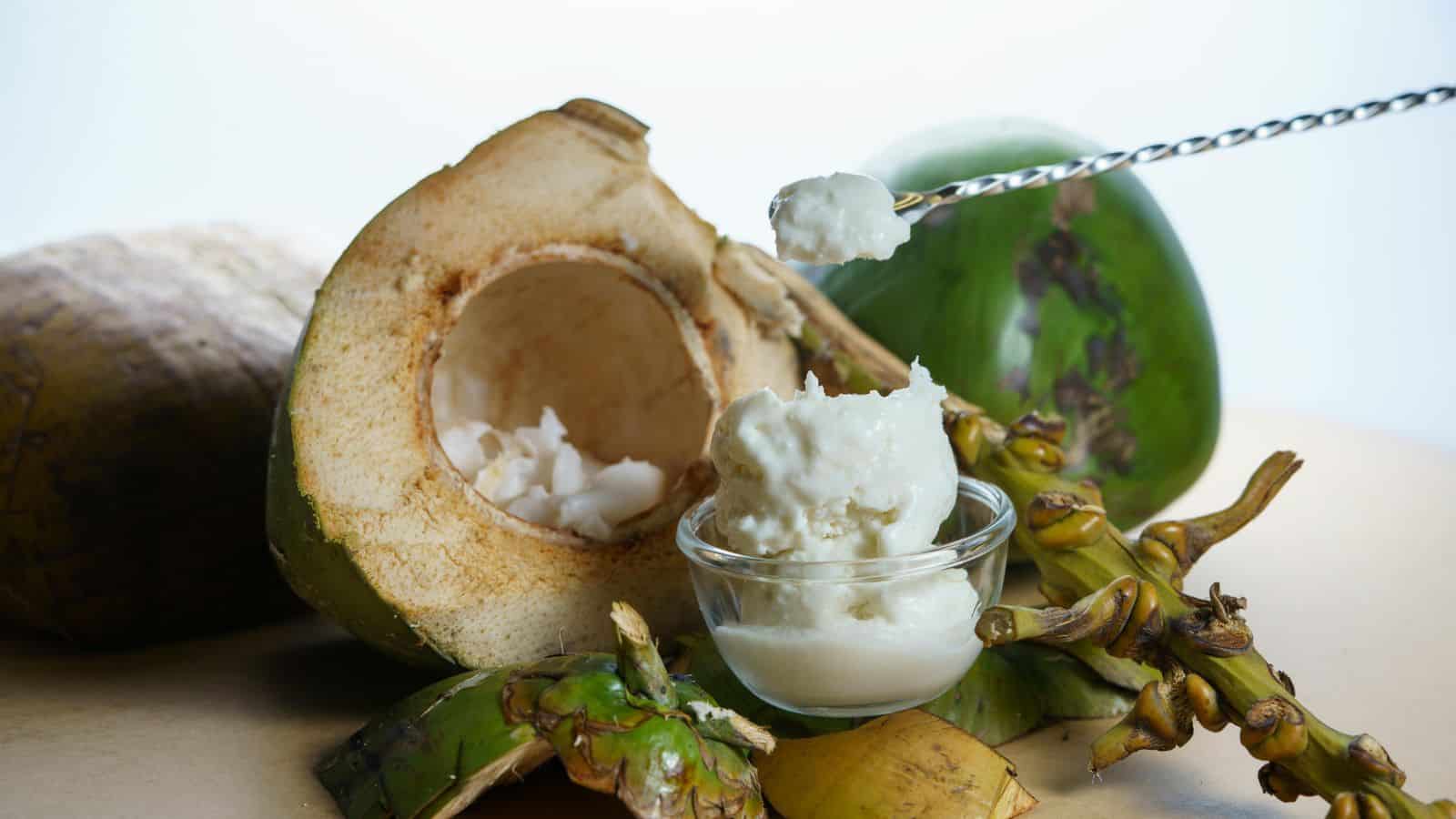 A split coconut with white flesh is displayed next to a small glass bowl filled with coconut cream. A metal skewer holds a dollop of the cream above the bowl. Green coconut husks and unopened coconut flowers are scattered around.