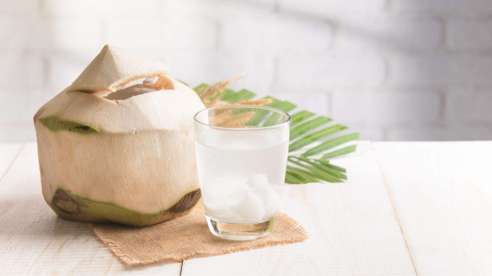 A fresh coconut with its top removed sits on a burlap cloth on a white wooden table. Next to it is a glass filled with coconut water and a few pieces of coconut flesh. A green palm leaf is partially visible in the background.