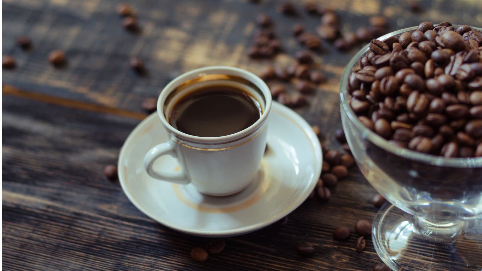 A white cup filled with black coffee sits on a saucer on a wooden table. Coffee beans are scattered around, and a glass container filled with coffee beans is partially visible on the right.