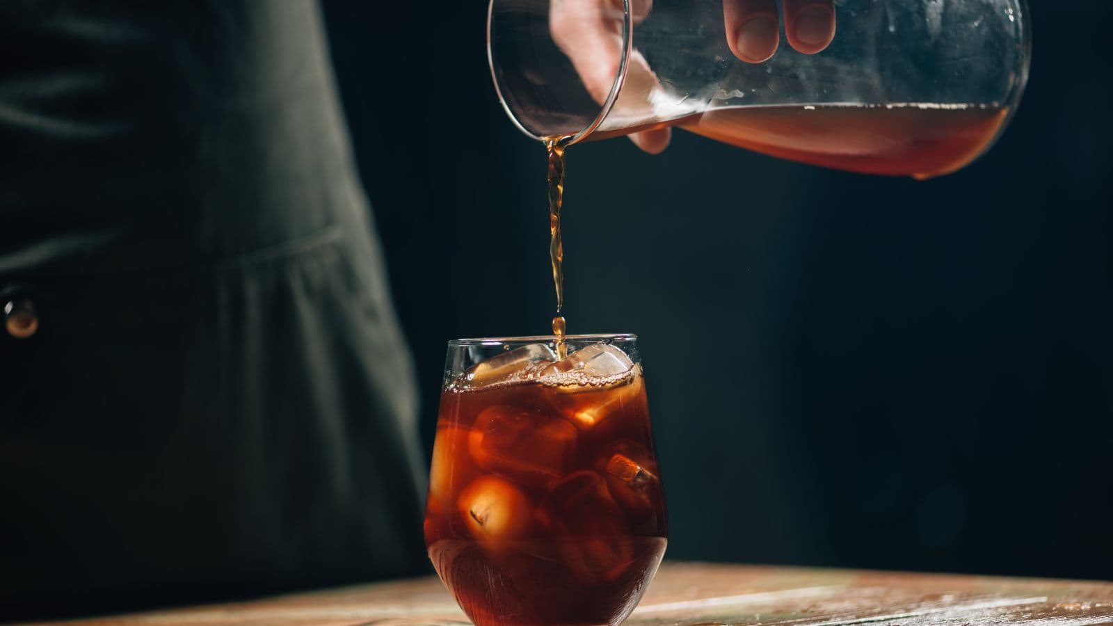 A hand is pouring cold brew coffee from a glass pitcher into a glass filled with ice. The glass is placed on a wooden surface, and the background is dark, highlighting the rich color of the coffee.