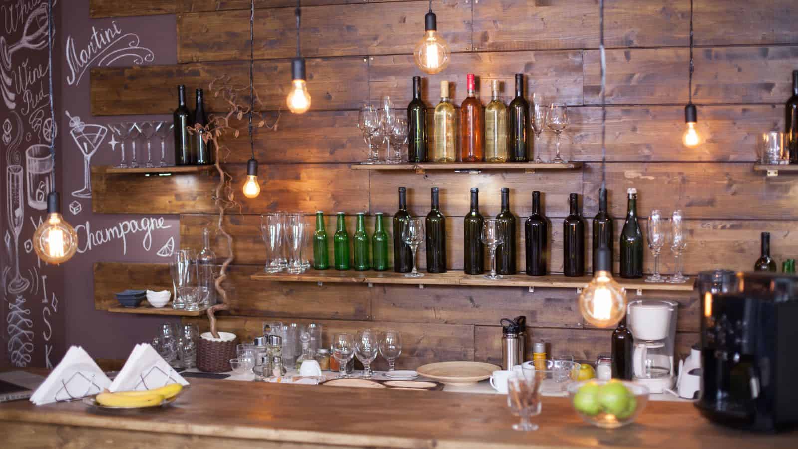 A rustic bar with wooden shelves adorned with various wine and liquor bottles. Hanging light bulbs provide warm lighting for this perfect holiday drink station. Chalkboard art decorates the wall, and glassware is neatly arranged on the counter, accompanied by a bowl of lemons.