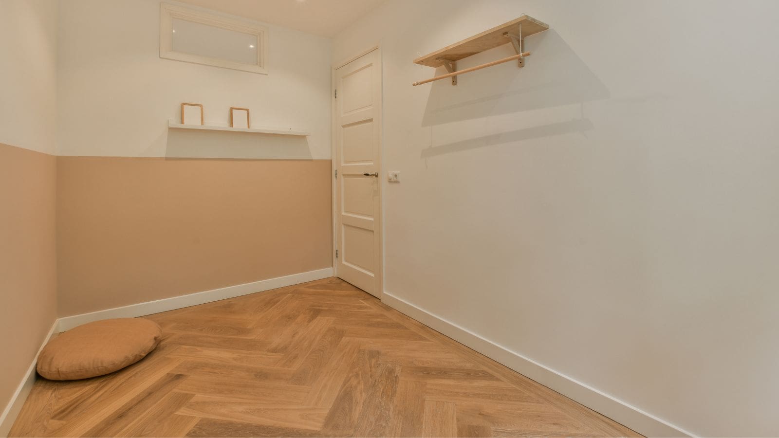A small, minimalist room with light wood flooring and beige walls. It features a closed white door, a wall shelf with two empty frames, and a round, brown cushion on the floor. There is a high window above the shelf.