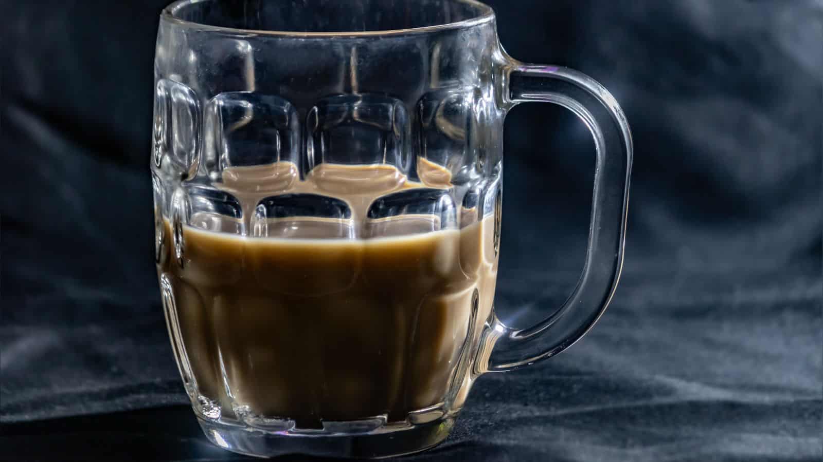 A glass mug filled with a dark brown liquid, possibly coffee, is placed on a dark, textured fabric background. The mug has a sturdy, classic design with a large handle.